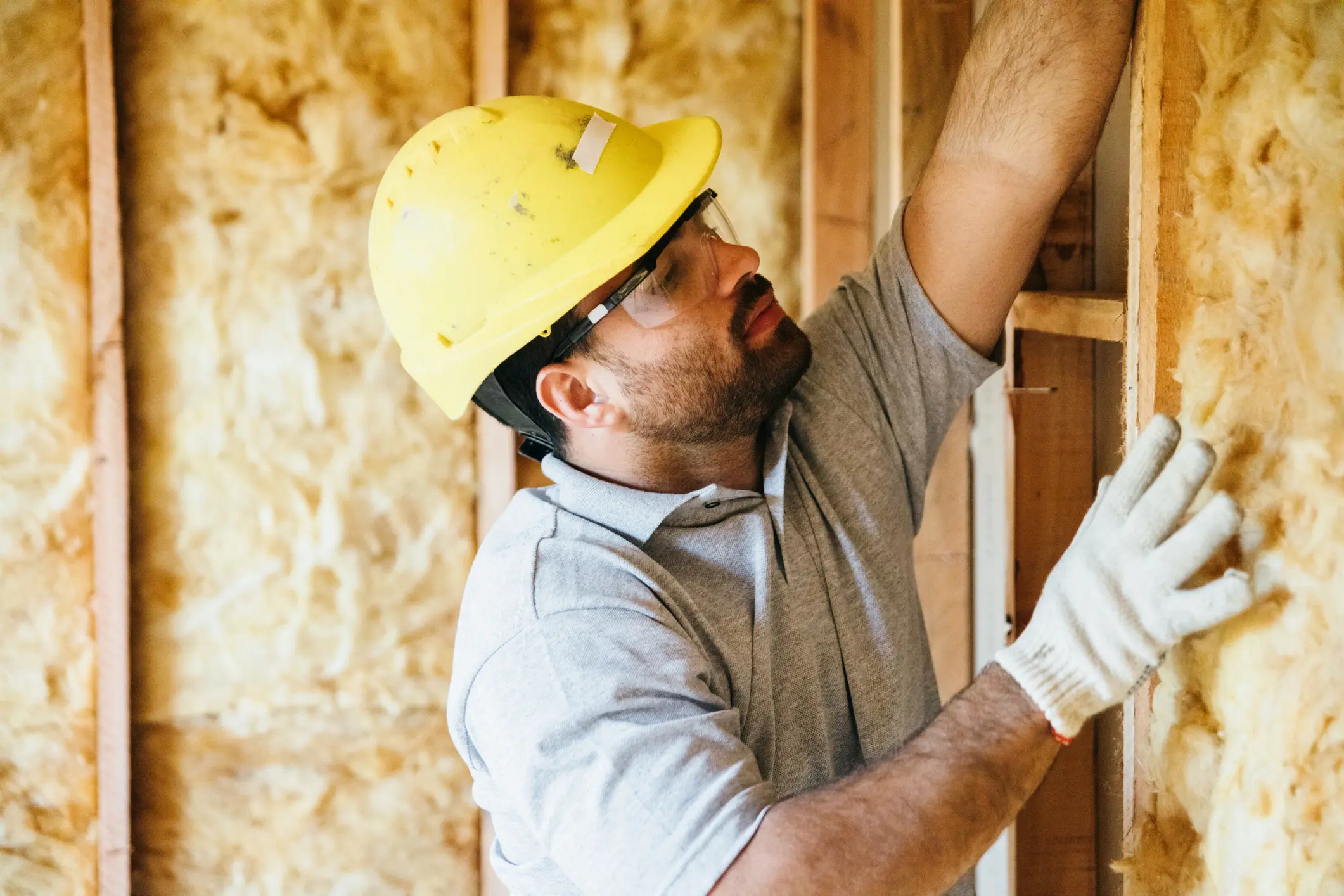 Worker in a hard hat and gloves professionally installing yellow fiberglass batt insulation between wall studs for residential home energy efficiency and comfort