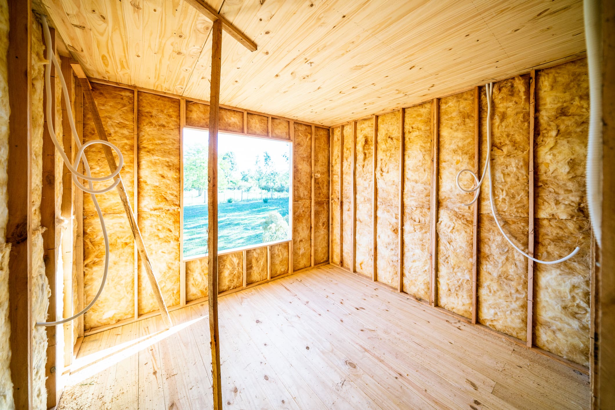 Room under construction with yellow fiberglass batt insulation installed between wooden studs and exposed pine ceiling and floor, sunlight through a large window overlooking green yard — home insulation, energy efficiency, spray foam alternative for Winona-area homeowners