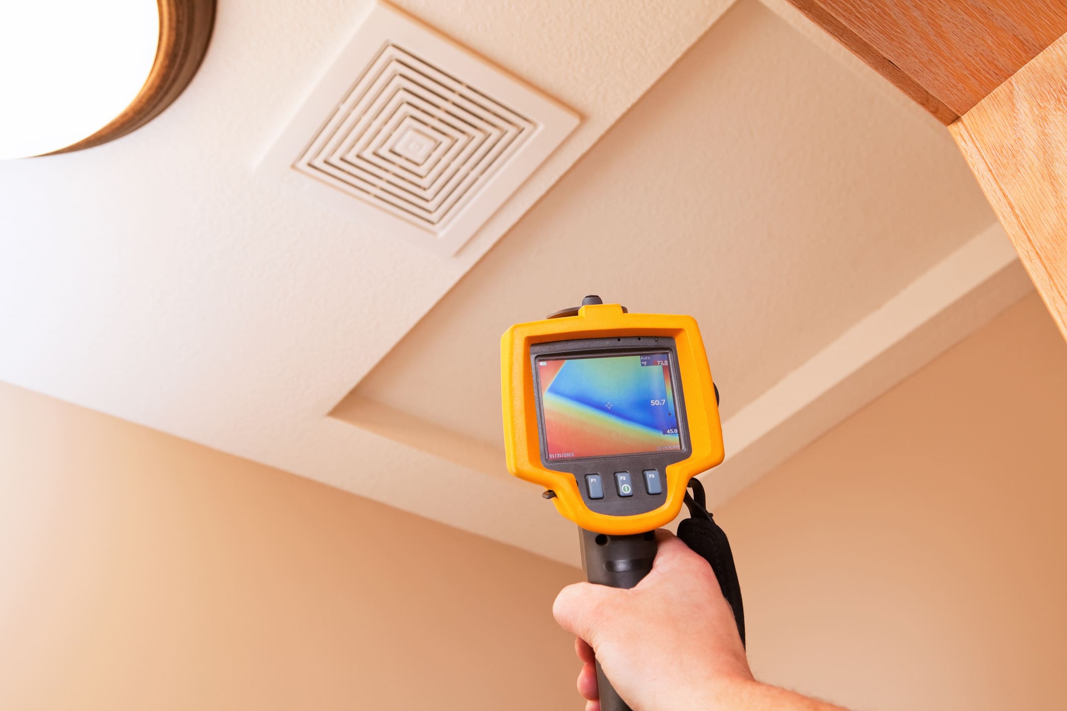 Hand-held thermal camera pointed at an attic access hatch and ceiling vent, showing a multicolor heat map used for attic insulation inspection and energy-efficiency assessment