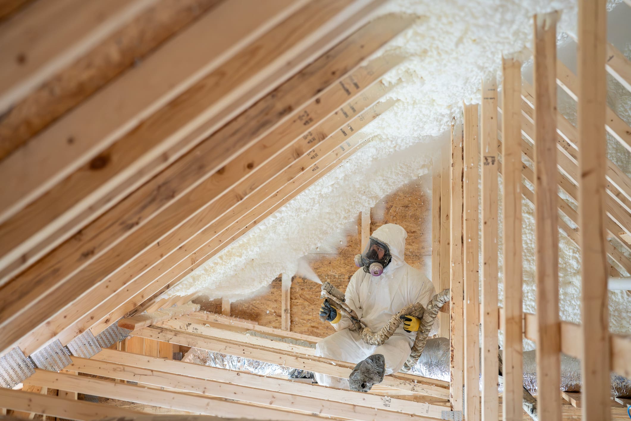 Contractor in full protective gear spraying white spray-foam insulation into an attic rafter cavity, illustrating A & H Insulation’s professional spray foam insulation installation for energy-efficient, comfortable homes and buildings